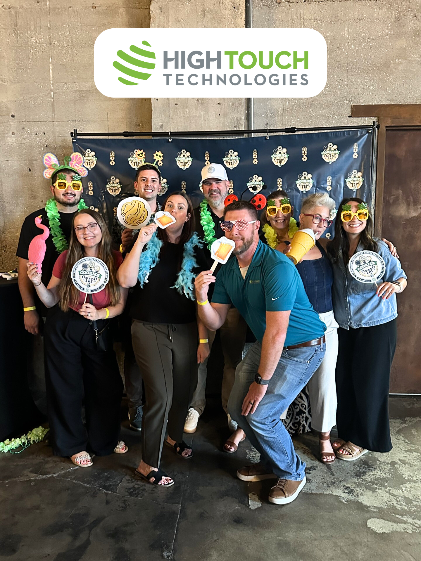 High Touch Technologies employees smiling and posing with fun props like sunglasses, flamingo cutouts, and cookie signs at the Cookies on Tap event photo booth.