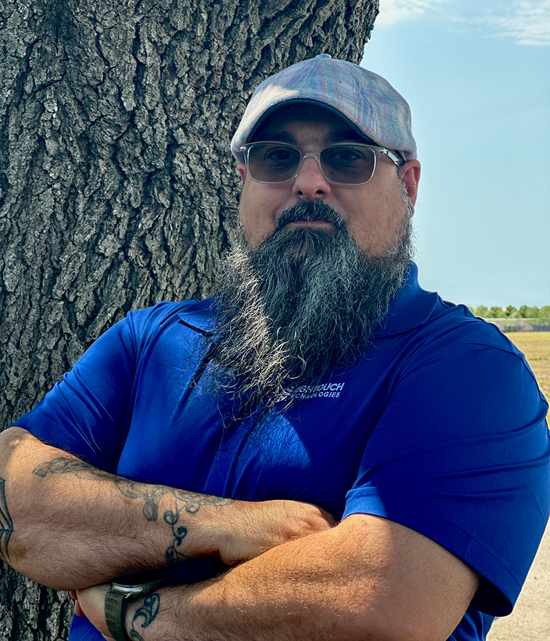 Josh Watkins, Systems Engineer II at High Touch Technologies, standing outdoors with arms crossed, wearing a blue company polo, flat cap, and glasses